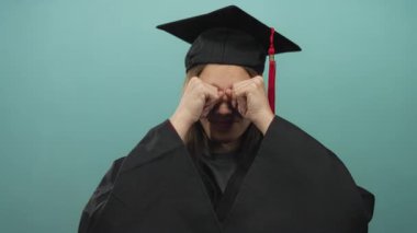 Mezuniyet kepi ve cübbeyle gözlerini ovuşturan genç bir kadın stüdyo içinde deniz mavisi arkaplanı ile; yorgunluk başarısı.