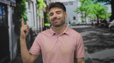 Adam güneşli şehir sokağında pembe polo tişört giyip kameraya gülümseyerek baş parmak hareketi yapıyor..