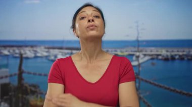 Kadın rıhtımdaki teknelerin önünde kollarını kavuşturuyor. Ötesinde görünen deniz olan bir binada çit var. İnkar ediyor..