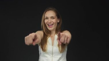 Siyah stüdyo ortamında iki işaret parmağıyla kamerayı işaret eden beyaz bir kadın..