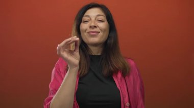 Hafif gülüşlü parlak pembe tişört giyen İspanyol kadın kırmızı stüdyoda yüzünün yanında işaret hareketi yapıyor..