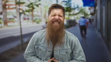 Kot ceket ve yeşil gömlek giyen sakallı genç adam, şehir caddesinde bulanık yayalarla karnını iki eliyle kavrıyor; acı.