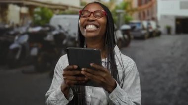 Şehir caddesinde elinde tabletle gülümseyen kırmızı gözlüklü ve çizgili bluzlu bir kadın şehir hayatını teknoloji ve mutlulukla sergiliyor..