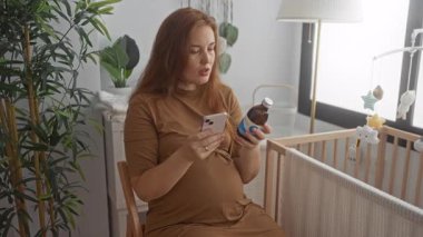 Bebek odasında karnının üzerinde akıllı telefon ve ilaç şişesi tutan hamile bir kadın. Annelik ve beklenti..