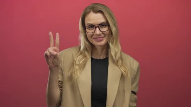 Kırmızı arka planda barış işaretleriyle poz veren sarı saçlı ve gözlüklü bir kadın..