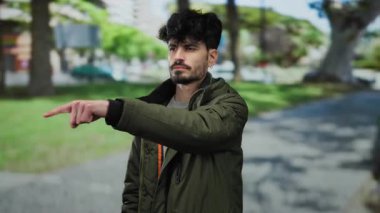 Sakallı genç adam, arka planda kararlılık ve odaklanma gösteren ağaçlar ve binalarla dolu bir parkta parkı gösteren bir ceket giyiyor..