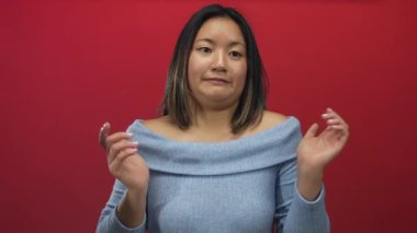 Kırmızı duvarın önünde omuz silken bir kadın, kafa karışıklığını ifade ediyor mavi kazak giyiyor ve dışa dönük bir hareket sergiliyor.