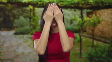 Kırmızı tişörtlü genç kadın, yeşilliklerle çevrili açık hava parkında neşeli bir şekilde duygularını ifade ediyor..