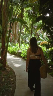 Lüks bir otelin verimli tropik bahçesinde yürüyen bir kadın elinde saman çantasıyla canlı yeşil yapraklarla sakin bir tatil ortamı yaratıyor.