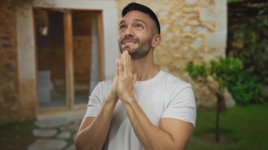 Beyaz gömlekli genç adam elleri dua ederken, taş duvarları ve yeşilliği olan kırsal bir Akdeniz evinin önünde gülümsüyor..