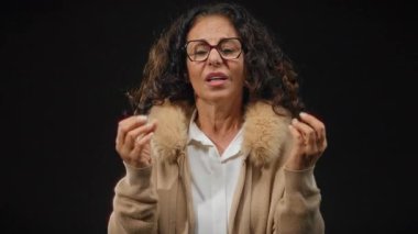 Kıvırcık saçlı ve gözlüklü İspanyol bir kadın. Siyah bir arkaplanda, kürklü bir palto giyiyor..