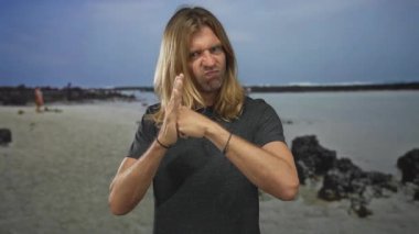Sarışın ve uzun saçlı bir adam, dalgaların çekildiği engebeli kayalık bir kıyı şeridinde avucuna parmağını bastırır; kararlılık.