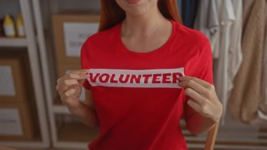 Bağış merkezinde kırmızı gönüllü gömleği sergileyen bir kadın. Arka planda malzeme rafları var. Kamu hizmeti ve destek ruhunu vurguluyor..