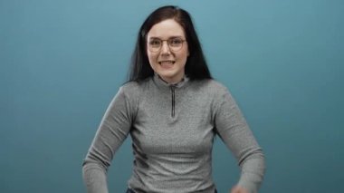 Esmer saçlı, mavi arka planda gözlüklü, farklı duyguları ifade eden ve gri kazak giyen bir kadın..