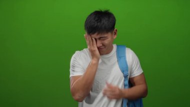 Beyaz gömlekli ve mavi sırt çantalı genç adam yeşil duvarın önünde duruyor. Stresli görünüyor ve yüzünü kapatıyor..