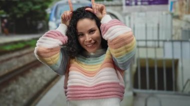 Renkli kazaklı genç İspanyol kadın açık bir günde arka planda canlı bir trenle açık hava tren istasyonunda şakacı boynuzlu jest yapıyor..