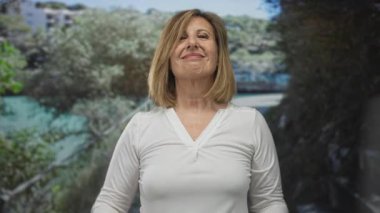 Ağaçlar ve güneş ışığıyla süslenmiş, beyaz gömlek giyen ve gülümseyen yüzlü, kendine güveni olan yeşil ormanda parmaklarını göğsüne gösteren bir kadın..