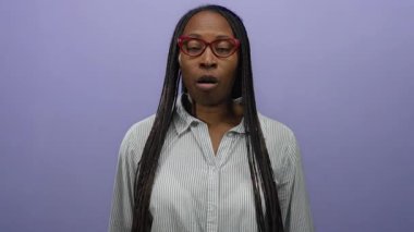 Gözlüklü, çizgili tişörtlü, esprili ve etkileyici kişilikli bir kadın, canlı mor bir arka plana karşı neşeli yüzler sergiliyor..