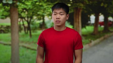 Çevresi yemyeşil ağaçlarla çevrili, açık hava parkı ortamında şüpheli görünen kırmızı gömlekli genç bir adam kendini sorgular ya da kimliğini açıklarmış gibi gösteriyor..