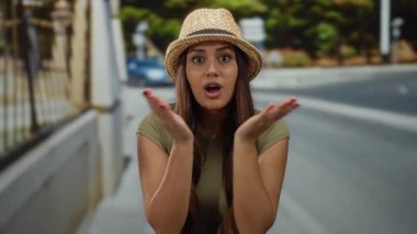 Şapkalı genç bir İspanyol kadın, elleri yüzü üzerinde bir caddede duruyor ve canlı bir şehir ortamında sürpriz yapıyor..