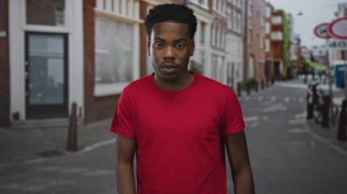 Kırmızı gömlekli adam işaret parmağıyla işaret ediyor ve işlek caddede tuğla binalarla yan bakıyor; merak.