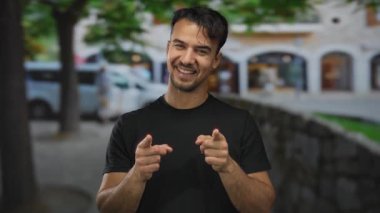 Genç İspanyol bir adam canlı bir şehirde neşeli bir şekilde dışarıyı işaret ediyor. Arka planda bulanık binalar ve ağaçlar var. Neşeli bir şehir yaşam tarzı sergiliyor.