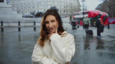 Yağmurlu bir günde şehir caddesinde gülümseyen bir kadın ikonik kırmızı bir otobüsle ve arka planda binayla neşeli bir şehir hayatı yaşıyor.