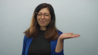 Mavi ceketli kadın gülümsüyor ve stüdyoda açık avuca işaret ediyor açık mavi arka planda; sıcak dostça bir davet.