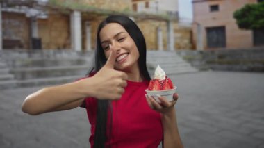 Dışarıda gülümseyen bir kadın bir kase çilekli dondurma tutuyor ve parmaklarını kaldırıyor, kırsal alanda kırmızı bir gömlek giyiyor..