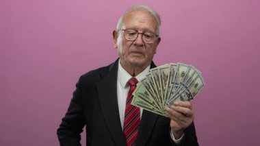 Takım elbiseli kıdemli bir adam elinde dolar banknotları ile pembe bir arka plana karşı, zenginliği ve iş başarısını odaklanmış bir ifadeyle temsil ediyor..