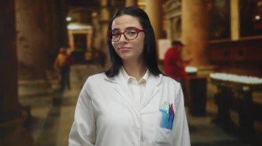 Beyaz üniformalı bir kadın, kırmızı gözlüklü, tarihi bir kilise arka planında, bilim ve mirası ön plana çıkarıyor..