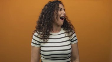 Kıvırcık saçlı ve ağzı açık İspanyol bir kadın stüdyoda turuncu arka plan önünde jest yapıyor. Sürpriz..