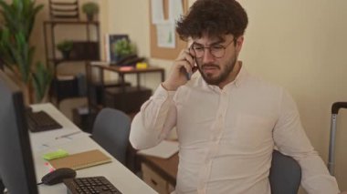 Ofiste sakallı genç adam telefonla konuşuyor. Masalar, bilgisayarlar ve bitkilerle çevrili. Bu da modern ve profesyonel bir çalışma alanı olduğunu gösteriyor..