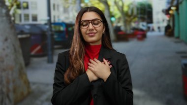 Gözleri kapalı, elleri yüreğinde gülümseyen bir kadın canlı bir şehir caddesinde kırmızı bluz ve siyah ceket giymiş, sıcakkanlı ve kendine güvenen bir kadın..