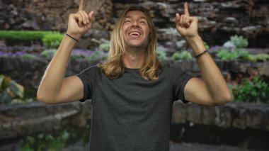 Uzun sarı saçlı, parmaklarını yukarı gösteren ve gün ışığında gür ormanda geniş bir gülümseme ile adam; sevinç merak.