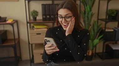 Modern ofis ortamında raflar ve bitkilerle akıllı telefon kullanan bir kadın gülümsüyor..