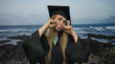 Mezuniyet üniformalı genç bir kadın sahilde neşe içinde ağlıyor. Arka planda deniz var. Açık deniz ortamında duygu ve kutlama karışımı bir şey sergiliyor..
