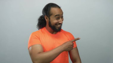 Açık gri stüdyoda, turuncu tişört giyen, göz alıcı ön kolu olan bir adam işaret ediyor..