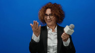 Esmer, takım elbiseli, mikrofonlu bir kadın, mavi arka planda heyecanla el kol hareketi yapıyor..