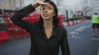 Şehir caddesinde inşaat çitlerinin yanında siyah gömlekli, alnından alnına kadar gölgelenen kadın, beklenti.