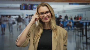 Havaalanında gözlüklü ve bej ceketli, düşünceli bir kadın kapalı bir terminalde dikilirken..