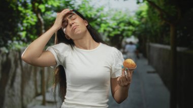 Yaz yeşilliğinin ortasında yapraklı ağaçlarla kaplı, güneşli bir sokakta kadın kek tutuyor ve alnına dokunuyor..