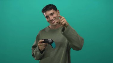 Latin asıllı genç adam elinde Cyan ve Black Game denetçisi gülümseyip yeşil stüdyoyu işaret ediyor; neşe dolu eğlence enerjisi.