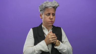 Mor stüdyoda elini göğsüne koyan kısa gümüş saçlı bir kadın ciddi bir ifadeyle; sessiz endişe yansıması.