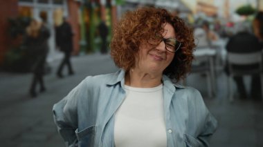 Kalabalık bir şehir caddesinde sırt ağrısı çeken bir kadın kıvırcık esmer saçlar, gözlük ve şehir hayatı ve açık havada oturma ile çevrili sıradan bir kıyafet sergiliyor..