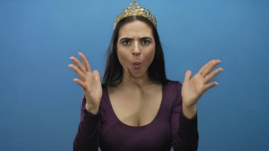 Mavi duvara taç takan genç İspanyol kadın neşeli bir şekilde elleri ve gözleri açık bir şekilde şaşkınlığını ve zevkini ifade ediyor.