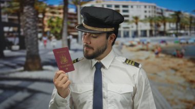 Sakallı pilot üniformalı genç adam açık mavi gökyüzünün altında deniz kenarında bir gezinti alanında Filipinli bir pasaport tutuyor..