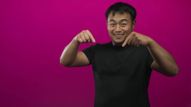 Siyah gömlekli gülümseyen adam boş alanı vurgulamak için pembe bir stüdyoda parmağını aşağı doğru gösteriyor; davet.