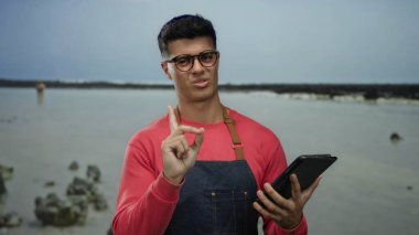Deniz kenarında tablet tutan önlük giyen bir adam merak gösteriyor. Sahilde düşünceli bir ifade ve arka planda kayalar var..