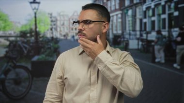Sokakta çeneye dokunurken bej gömlek giyen ve gözlük takan İspanyol bir adam..